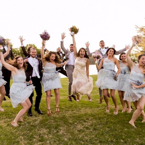 A joyful wedding party jumps in the air, with bridesmaids in light blue dresses and groomsmen in suits, celebrating outdoors on a sunny day.