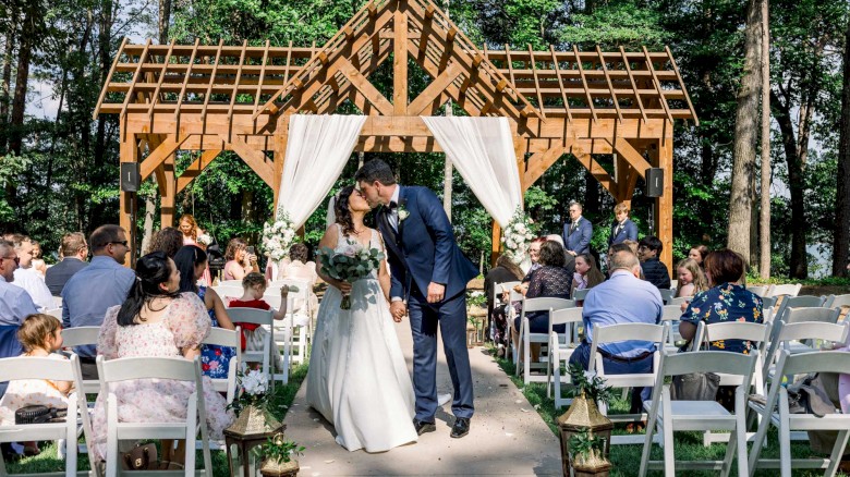 A couple is kissing at their outdoor wedding ceremony under a wooden arbor, surrounded by guests seated on white chairs in a lush, green setting.