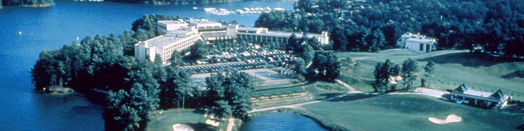 An aerial view of a lakeside resort complex featuring a large building, surrounding wooded areas, a golf course, and multiple water bodies ending the sentence.