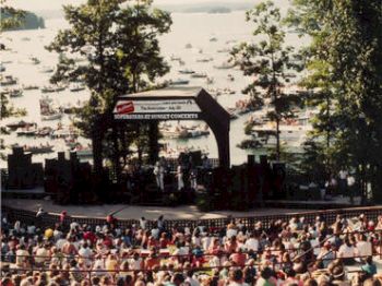 The image shows an outdoor concert by a lake, with a crowd watching from a seated area and numerous boats on the lake in the background, ending the sentence.