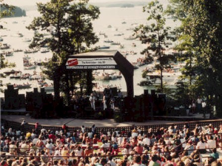 The image shows an outdoor concert by a lake, with a crowd watching from a seated area and numerous boats on the lake in the background, ending the sentence.