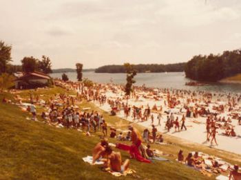 A crowded beach scene with numerous people sunbathing and swimming near a lake with a grassy hill in the foreground ending the sentence.
