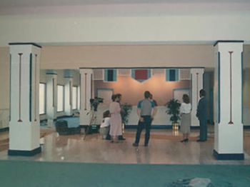 The image shows a group of people in a lobby-like room with pillars and plants. The group appears to be in discussion or an interview setup.