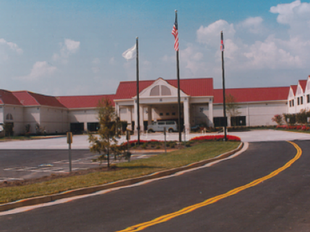 A building with red roofing and multiple flags in front, surrounded by an asphalt driveway and some landscaping, on a clear day.