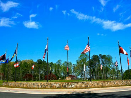 A circular monument with multiple national and state flags surrounded by a stone base and greenery under a clear blue sky with scattered clouds.