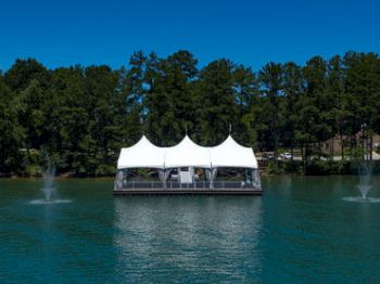 A floating structure with a white tent stands on a serene lake, surrounded by trees, with two water fountains in the foreground.