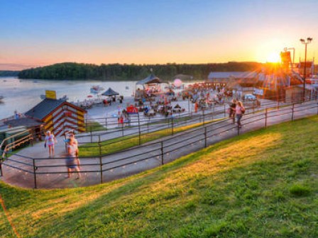 An outdoor scene with people walking along a path on a grassy hill, next to a body of water with tents, tables, and lights during sunset.