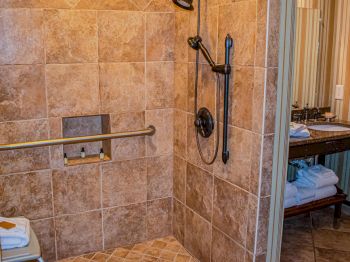 This image shows a tiled shower with a grab bar, handheld showerhead, and built-in shelf, adjacent to a sink area with towels and toiletries.
