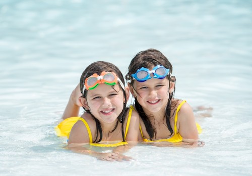 Two children in yellow swimsuits with goggles are smiling while lying in the shallow water at a pool or beach.
