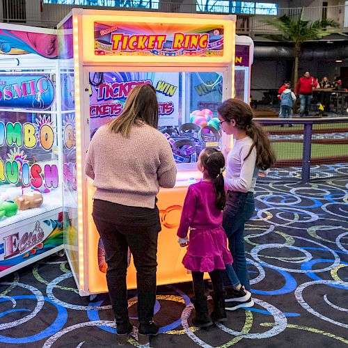 Three people, including two children, are standing by an arcade crane machine in an entertainment area with colorful flooring and other arcade games.
