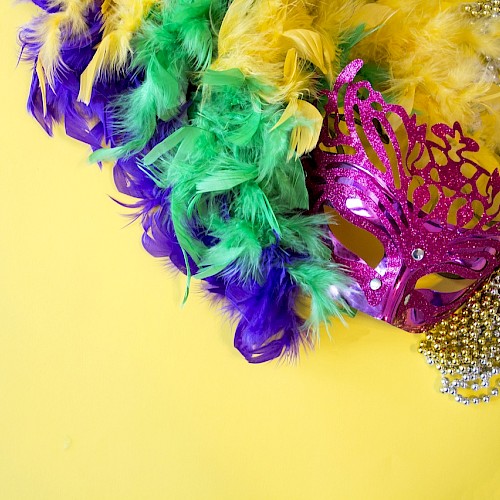 A pink masquerade mask with green, yellow, and purple feathers, and a string of gold beads on a yellow background.