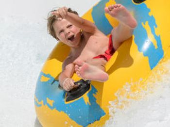 A child is joyfully riding a yellow and blue inflatable tube down a water slide, splashing water around with a big smile and raised arms.