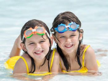 Two children wearing yellow swimsuits and goggles are lying in shallow water, smiling.