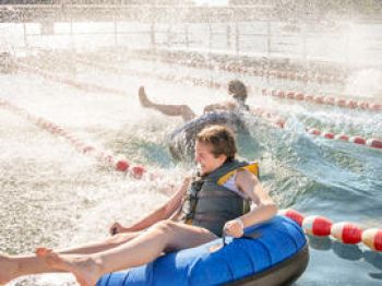Two people enjoying water tubing in an outdoor pool, one on a blue inflatable tube splashing water everywhere.