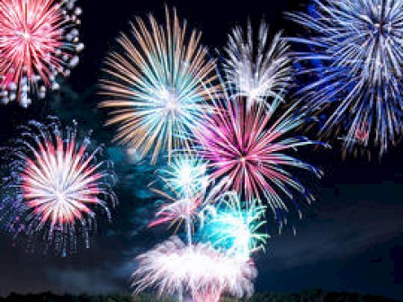 The image shows a vibrant display of fireworks lighting up the night sky over a body of water, reflecting the colorful explosions.