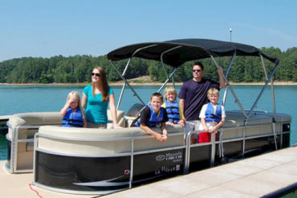 A family is on a pontoon boat docked at a lake; everyone is smiling and some are wearing life jackets while enjoying a sunny day.