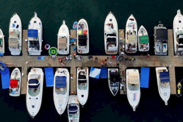 An overhead view of a marina with several boats docked along a pier, surrounded by people and various equipment on and around the boats.