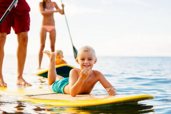 A family is paddleboarding on the water; a child is lying on a yellow board smiling, while another person paddles in the background.