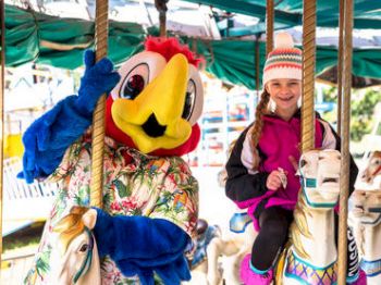 A person in a colorful bird costume and a girl wearing a beanie hat and jacket are riding carousel horses, smiling at the camera.