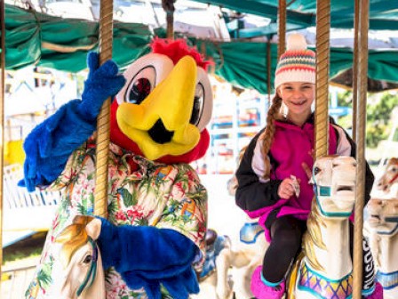 A person in a colorful bird costume and a girl wearing a beanie hat and jacket are riding carousel horses, smiling at the camera.