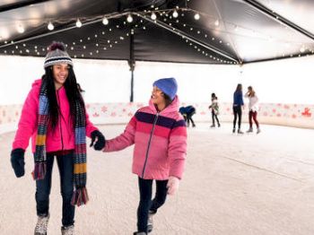 People are ice skating under a decorated tent. Two individuals in colorful winter clothing are holding hands and smiling, enjoying the activity.