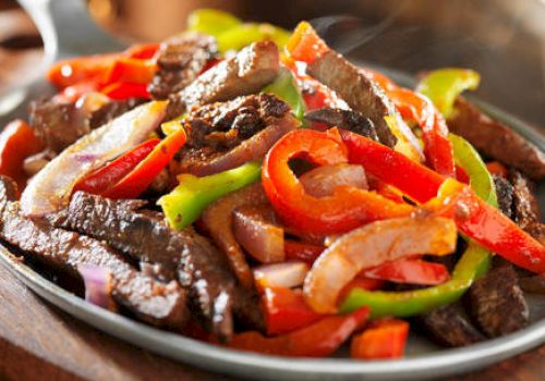 The image shows a dish of sizzling beef fajitas with colorful bell peppers and onions served on a hot skillet on a wooden board.