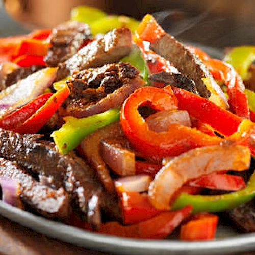 The image shows a dish of sizzling beef fajitas with colorful bell peppers and onions served on a hot skillet on a wooden board.