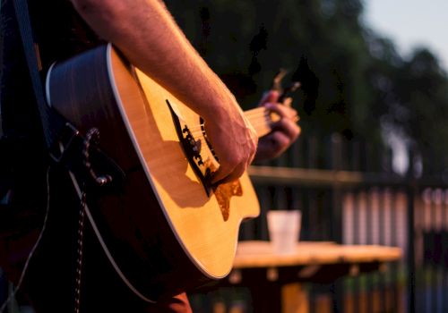 A person is playing an acoustic guitar outdoors, with a table and a drink in the background. The focus is on the guitar and the musician's hands.