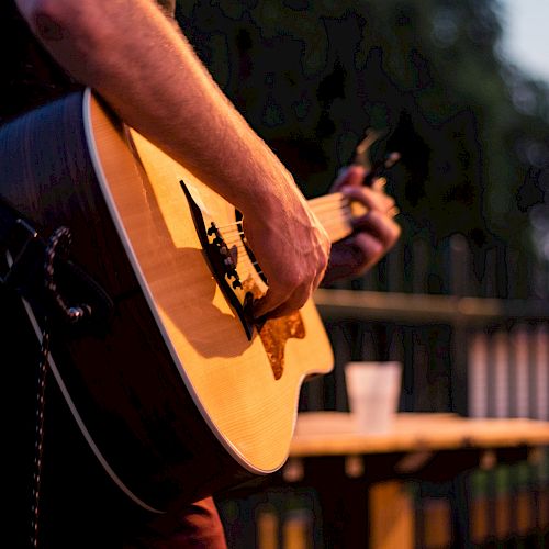 A person is playing an acoustic guitar outdoors, with a table and a drink in the background. The focus is on the guitar and the musician's hands.