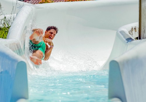 A person is sliding down a water slide, splashing water, and appearing excited or having fun. The slide has white sides and a water pool below.