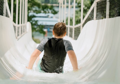A child is sliding down a water slide, wearing a dark shirt, with arms extended. The slide has net sides, and the background is blurred greenery.