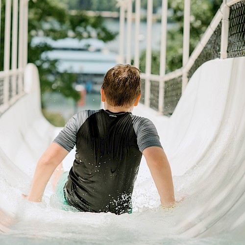 A child is sliding down a water slide, wearing a dark shirt, with arms extended. The slide has net sides, and the background is blurred greenery.