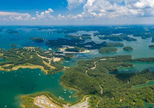 This image shows an aerial view of a lake featuring many small islands, boats, and surrounding greenery under a partly cloudy sky.