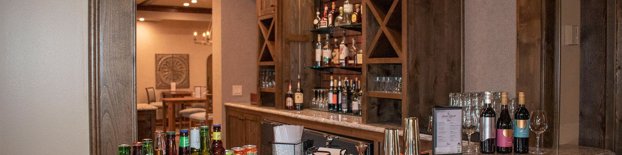 This image shows a home bar with bottles of beer, wine, and liquor on a granite countertop, wooden cabinets, and a view into another room.