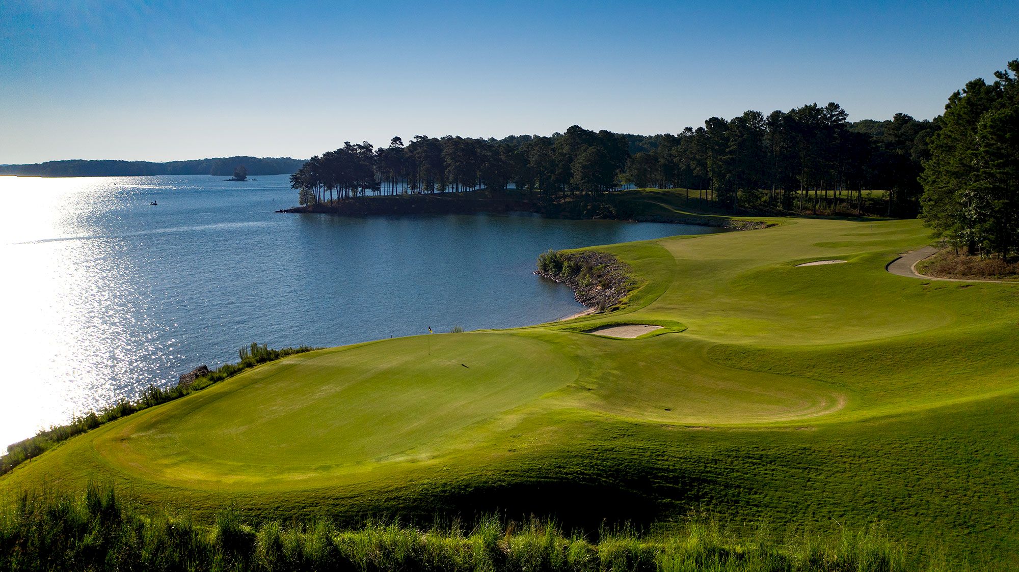 This image shows a serene golf course by a body of water with lush greenery, sand traps, trees, and a clear blue sky in the background.