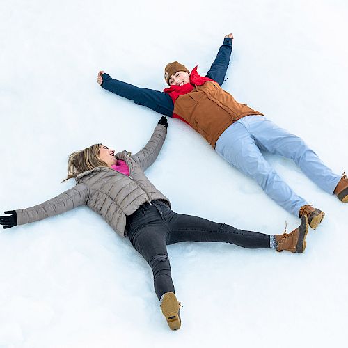 Two people are lying on the snow, making snow angels with their arms and legs spread out, dressed warmly in jackets, hats, and boots.