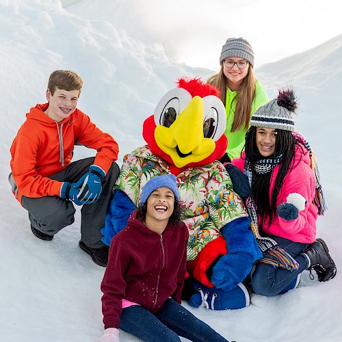 Four people and a bird mascot are posing in the snow, all smiling and wearing winter clothes.