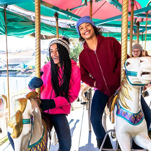 Two people are riding a carousel, smiling and wearing winter clothes. Other people are visible in the background, also enjoying the ride.