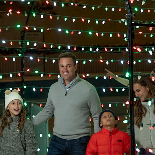 A family is enjoying the holiday lights, with a man and woman walking with two children under colorful festive lights, smiling and happy.