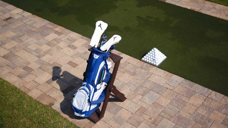 A golf bag with clubs is placed on a stand next to a pile of golf balls on a small grassy practice area with a brick-patterned surface.