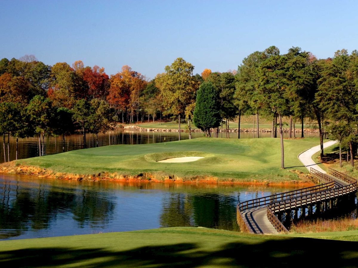 The image shows a golf course featuring a green surrounded by water, with a footbridge and a background of trees displaying autumn colors, ending the sentence.