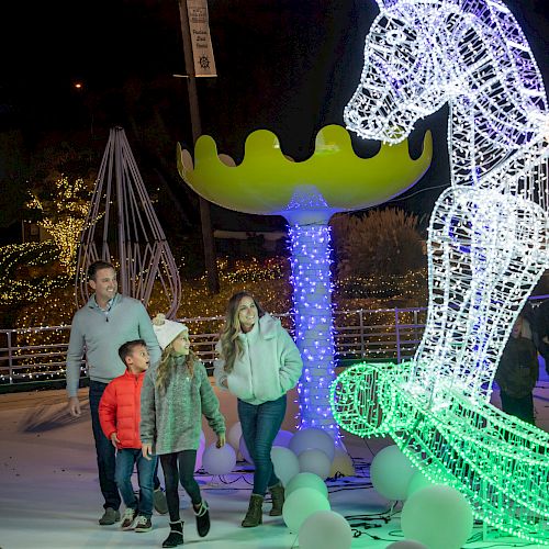 A family walks through a festive light display featuring a large illuminated rocking horse and other decorative elements at night.