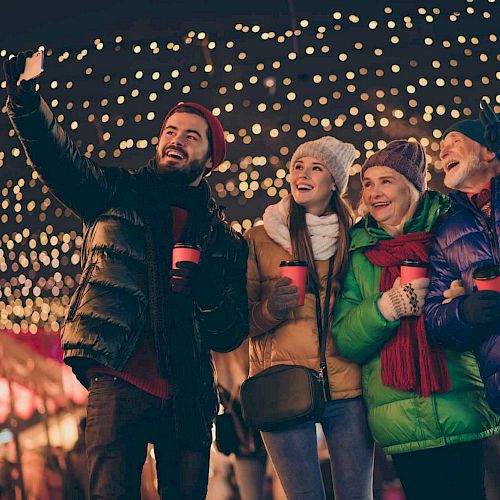 A group of four people, wearing winter clothing, are enjoying a festive outdoor setting with string lights, smiling, and taking a selfie.