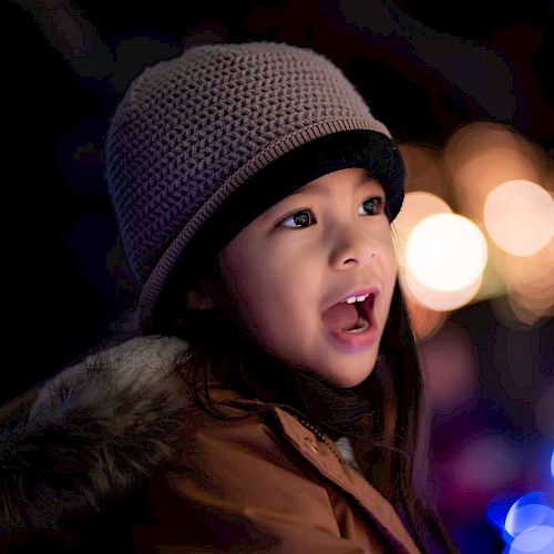 A child wearing a hat and jacket is looking excitedly at something, with colorful, out-of-focus lights in the background creating a festive atmosphere.