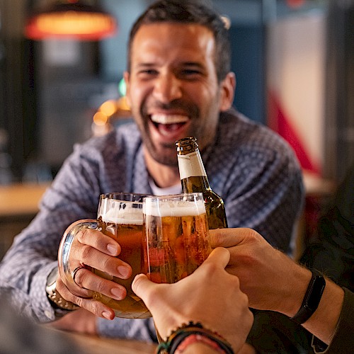 Three friends are enjoying drinks together at a bar, smiling and laughing while toasting with beer mugs and a bottle.