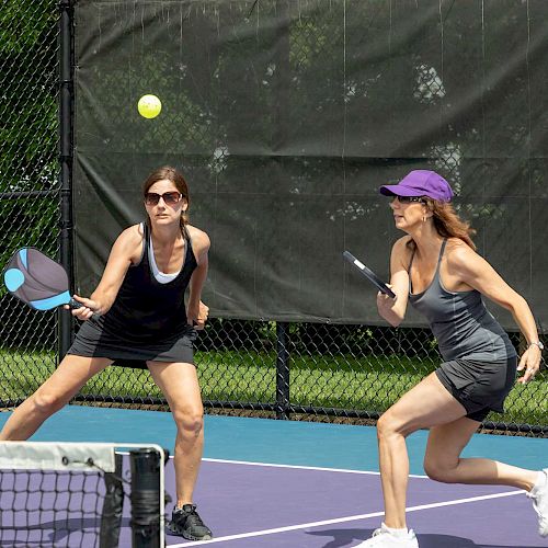 Two women are playing pickleball on an outdoor court, both focused on an incoming ball.