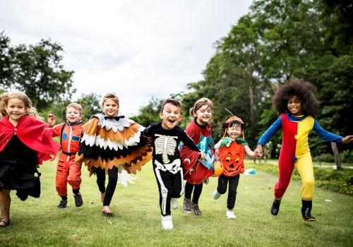 A group of children in various costumes, including a skeleton, a pumpkin, and a bird, are joyfully running on a grassy area outdoors, ending the sentence.