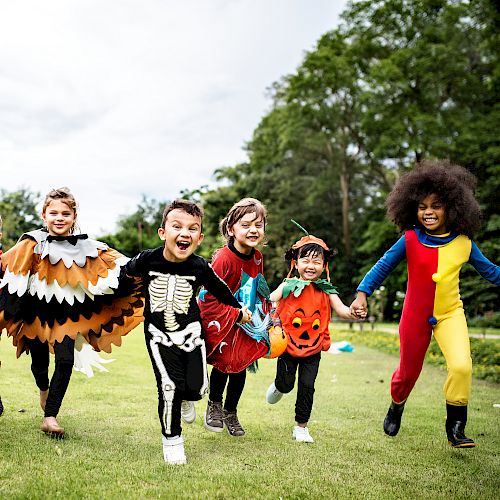 A group of children in various costumes, including a skeleton, a pumpkin, and a bird, are joyfully running on a grassy area outdoors, ending the sentence.