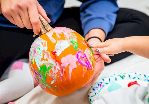 Two people are painting a pumpkin with colorful paints, creating a vibrant design together.