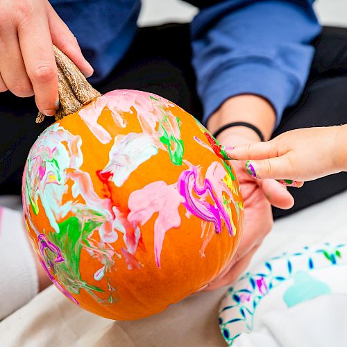 Two people are painting a pumpkin with colorful paints, creating a vibrant design together.